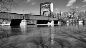 Mud pulls at your feet, the river hums under closed bridges, and the air thickens with thawed-out memory. Between branches and broken trails, a stillness opens—where water dreams upward, steel waits without speaking, and the season writes itself in soft collisions.