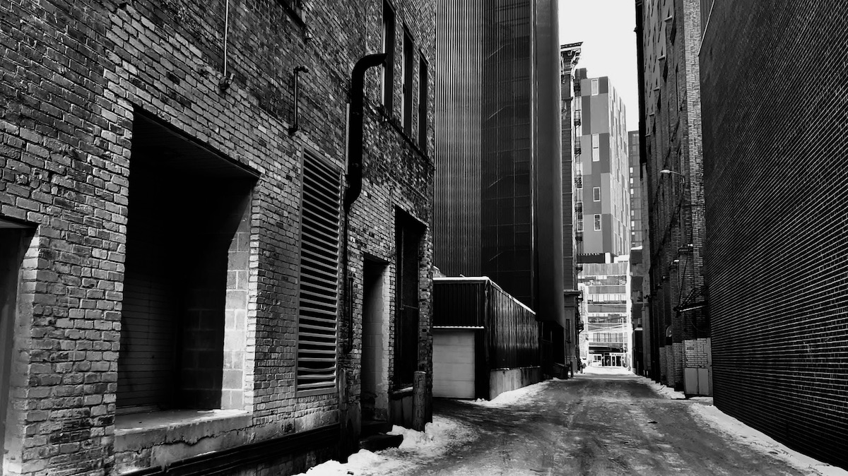 This Winnipeg alley dreams in grayscale, where nothing begins and nothing quite ends. It folds space like a tired map, worn soft at the creases. Light doesn’t fall here—it drifts, uncertain, like memory losing its edges. Brick holds the breath of things unsaid, while the ground carries a quiet ache, neither sorrow nor peace. The buildings don’t lean—they hover, caught between presence and forgetting. In this suspended stillness, time exhales slowly, then disappears, leaving behind only the echo of having once been.