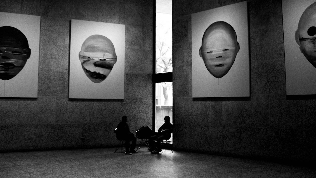 They gather beneath giants—paint, steel, and silence pressing down like weather. Here, they are not exhibits but echoes, resisting the stillness with their own weight of being. This is not interruption. This is grounding. A reminder that presence is also a kind of art.