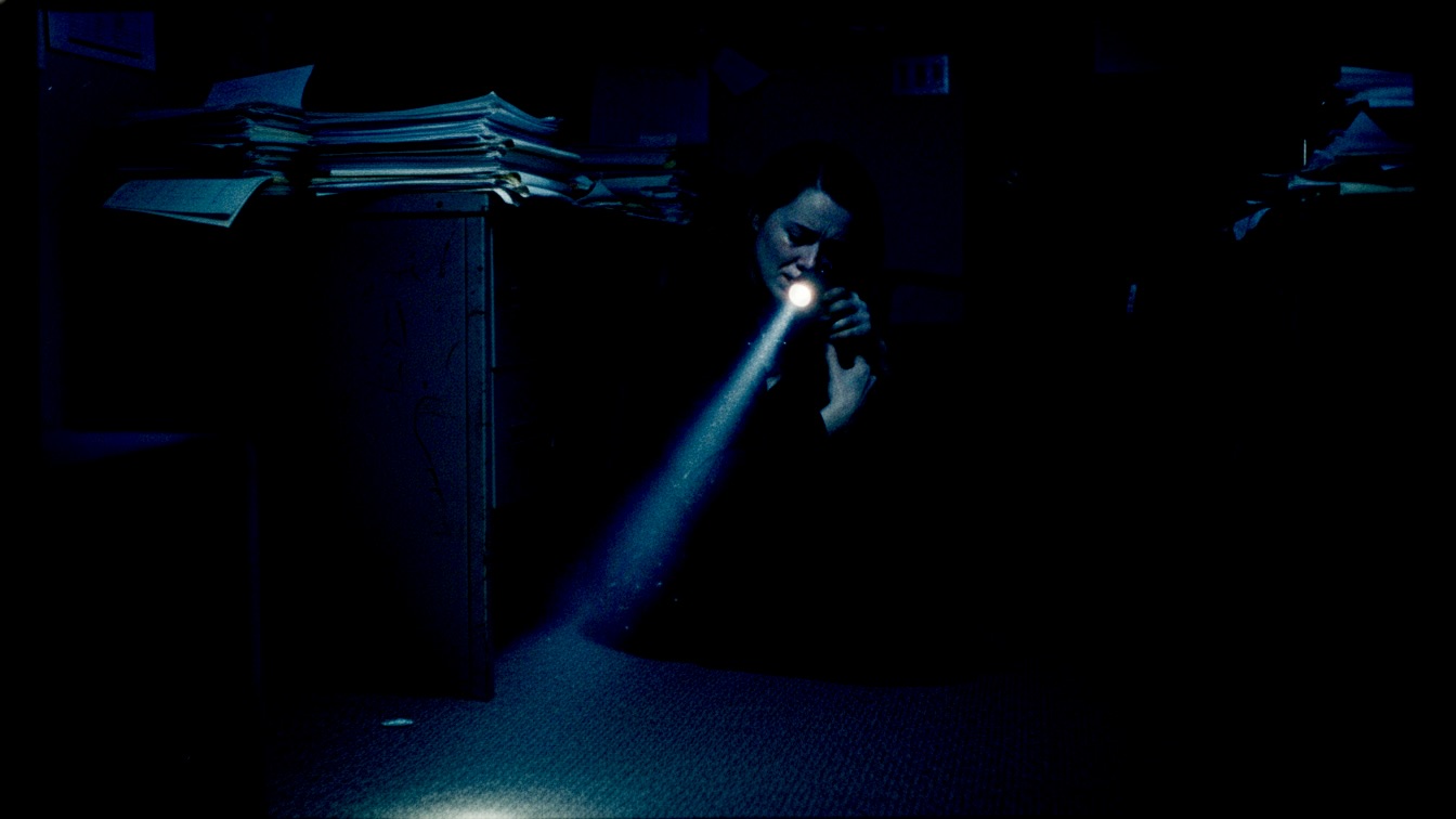 A flashlight beam illuminates a woman huddled in a dark office while a man sits in shadow.