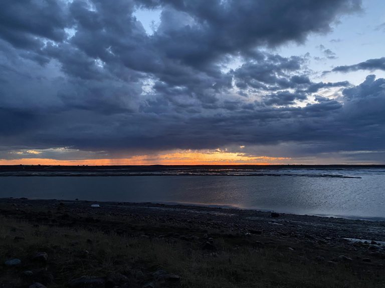 The sun begins its descent, casting a golden hue over the serene waters and the rugged terrain. The sky is painted in shades of orange and pink, highlighting the tranquil yet vibrant atmosphere of the coming night across the western shores. Photo: Tony Eetak