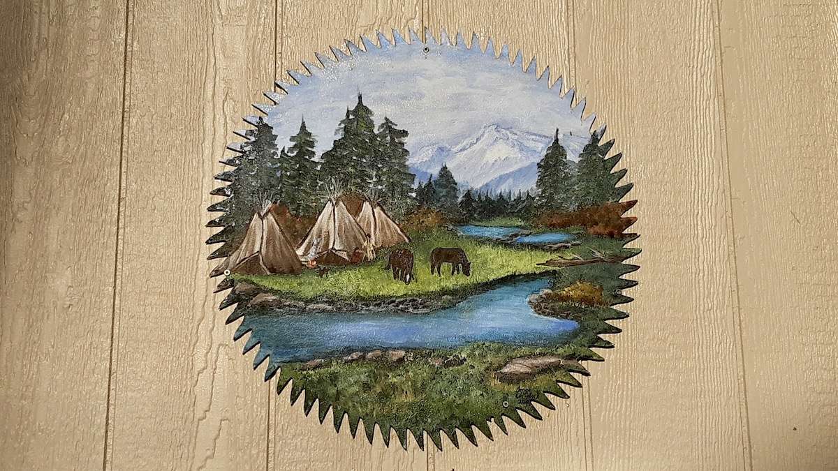 The jagged edges of a saw blade frame a tranquil landscape of mountains, forests, and rivers in this unique artwork by Tanya McArthur from Dryden.