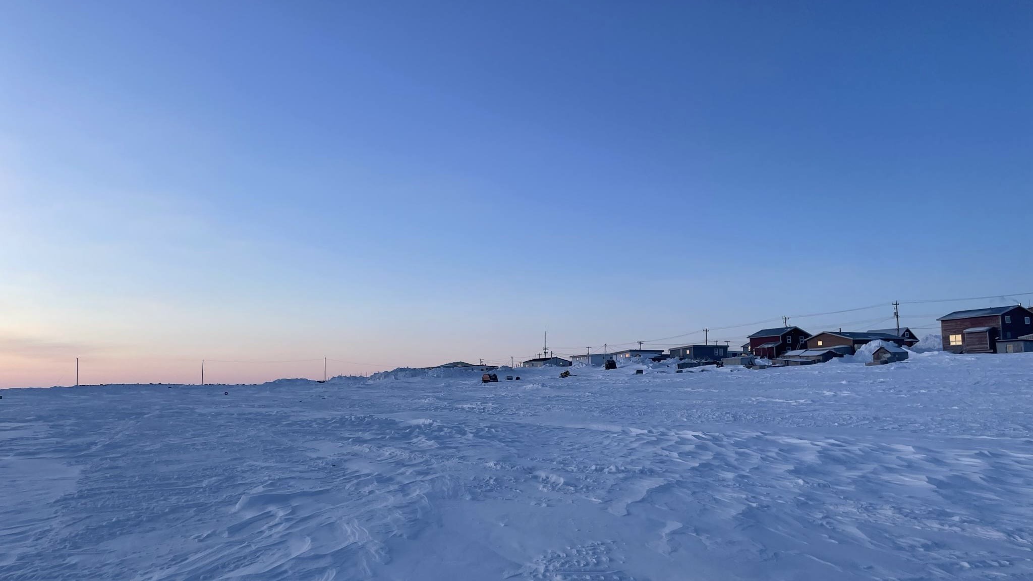 Sky and snow, meeting our homes. A soft light touches everything. Familiar shapes, but a different feel in the air. The edge of our community, where the wild meets our lives. We watch, we remember, we see the subtle shifts in the world around our doors. This is our place, changing with the light.