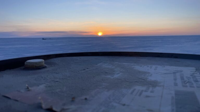 A metal drum, dusted and fading, frames the edge of land and light. The sun breaks clean across an ice-locked horizon, distant yet grounding. Industrial memory sits in the foreground, silent but loaded. This is the balance: between use and loss, sky and surface, heat and cold, stillness and what is coming next.