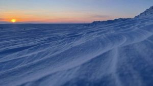 The setting sun paints the snow-covered shores of Arviat with soft hues where the frozen expanse of Hudson Bay meets the land. Wind-sculpted drifts create a stark beauty under the fading light, a silent testament to the Arctic winter giving way to the subtle promise of spring.