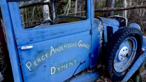 This weathered side panel, likely belonging to a Ford Model A or similar early 20th-century vehicle, features the ghosted hand-painted signage of "Percy Anderson's Garage Dyment." The bold lettering, despite decades of exposure, serves as a tangible link to a local business from a bygone era.