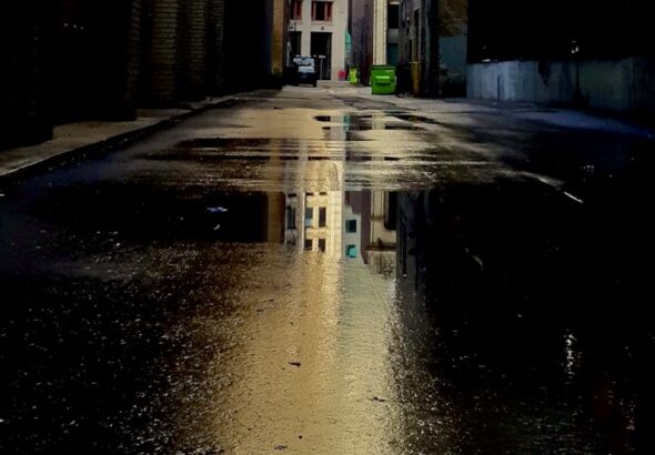 Rainwater pools on the worn pavement of a downtown Winnipeg alley, creating a striking mirror that reflects historic architecture and invites late-night walkers to contemplate a century of urban change.